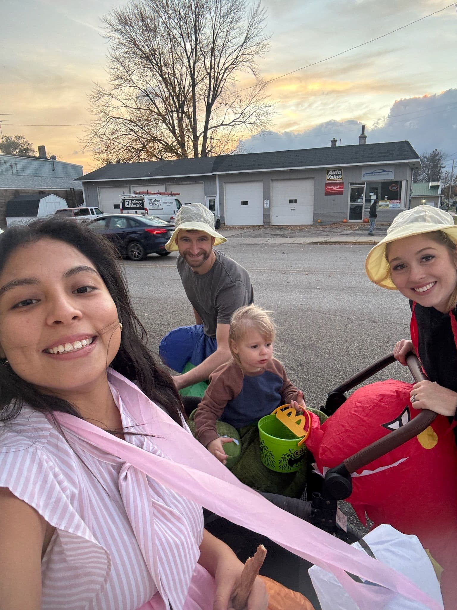 Selfie grupal en un atardecer de Halloween. Tres adultos y un niño con disfraces y cubetas de dulces frente a un taller.